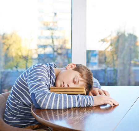 Sleepy 12 years old children boy sitting at the wooden desk and sleeping over the book, composition against the windowの写真素材