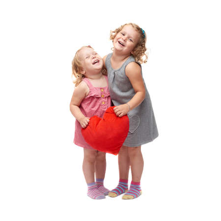 Couple of young little girls sisters with curly hair in gray and pink dress holding red plush heart and standing over isolated white backgroundの写真素材