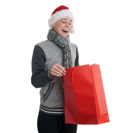 Cute teenager boy in gray sweater and christmas hat looking into red shopping bag over white isolated background, half bodyの写真素材