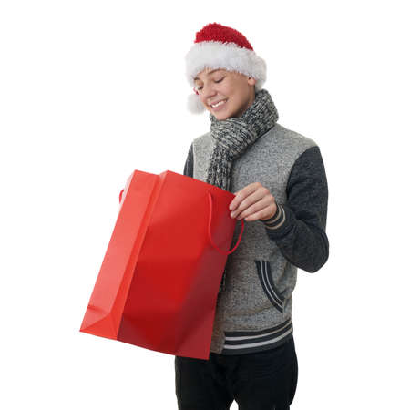 Cute teenager boy in gray sweater and christmas hat looking into red shopping bag over white isolated background, half bodyの写真素材