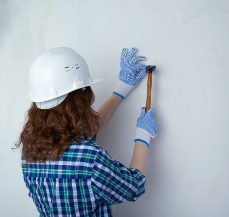Young woman in casual clothes in front of white unpainted wall in white helmet working with hammer, happy people and construction conceptの写真素材