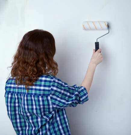 Young woman in casual clothes in front of white unpainted wall working with paint roller, happy people and construction conceptの写真素材