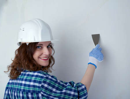 Smiling young woman in casual clothes in front of white unpainted wall in white helmet working with putty knife, happy people and construction conceptの写真素材