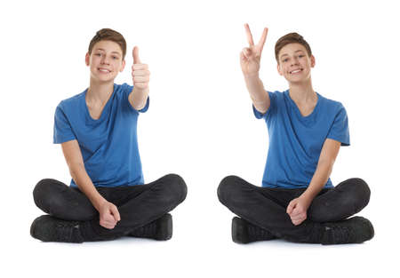 Cute teenager boy showing thumb up victory sign in blue T-shirt and lotus posture over white isolated backgroundの写真素材