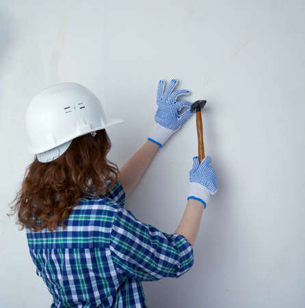 Young woman in casual clothes in front of white unpainted wall in white helmet working with hammer, happy people and construction conceptの写真素材