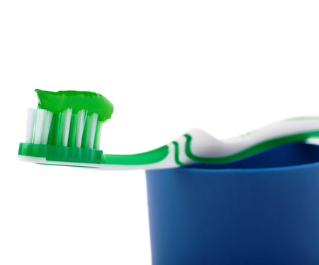 Green single toothbrush with paste lying on blue plastic cup isolated over white background