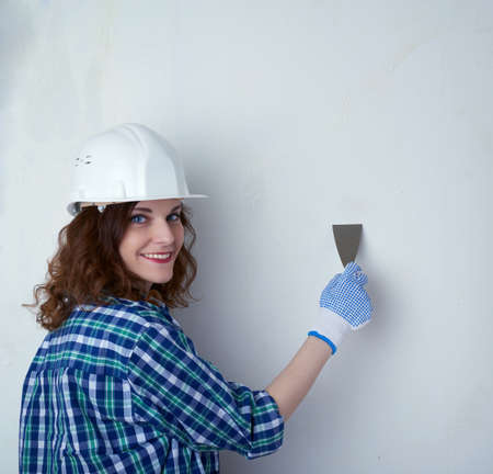 Smiling young woman in casual clothes in front of white unpainted wall in white helmet working with putty knife, happy people and construction conceptの写真素材
