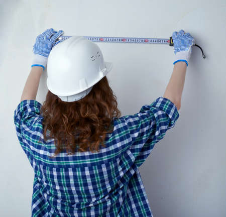Young woman in casual clothes in front of white unpainted wall in white helmet working with measuring tape, happy people and construction conceptの写真素材
