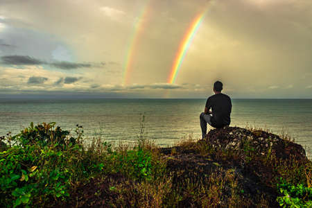 man at hilltop enjoying amazing landscape with rainbow above sea horizon image is taken at gokarna karnataka india. the view from here is pristine and mesmerizing.の写真素材