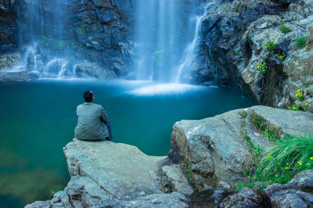 young man watching waterfall falling from mountain with calm blurred water surface at morning image is taken at thangsning fall shillong meghalaya india.の写真素材