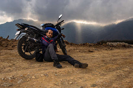 man sitting near motorcycle at mountain top with dramatic sky at eveningの写真素材
