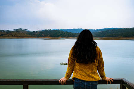 young girl looking the serene natural beauty at lake shore from flat angle image is taken at umiam lake shillong meghalaya india.の写真素材