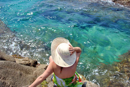 woman sitting on sea rocks looking at the horizon with his hatの写真素材