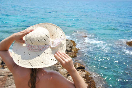 woman sitting on sea rocks looking at the horizon with his hatの写真素材