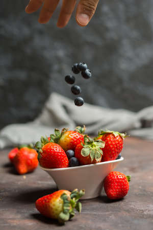 Full of ceramic bowl with blueberries and strawberries  on the stone table. And a hand dropping pieces of blueberries from the airの写真素材