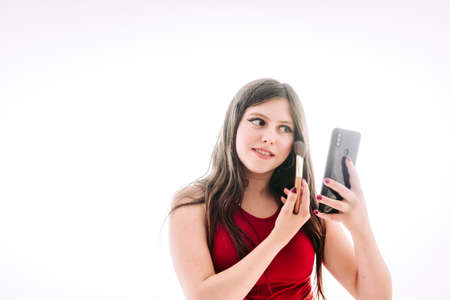 Teenage girl putting on a little makeup, seeing herself reflected on the screen of her mobile phone.の写真素材