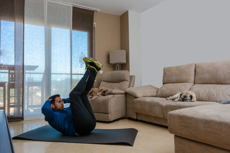 Latin man, doing a workout in his living room, does sit-ups, stretches and squatsの写真素材