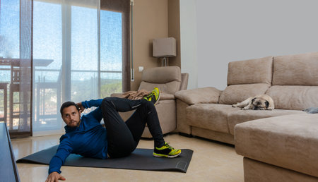Latin man, doing a workout in his living room, does sit-ups, stretches and squatsの写真素材