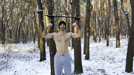 Portrait of young shirtless sportsman standing in snowy forest and showing biceps. Handsome strong...