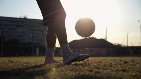 Legs of young man kicking ball at green field. Male feet of professional footballer juggling soccer ball on stadium at sunset. Sportsman practicing tricks outdoor. Concept of a freestyle footballの写真素材