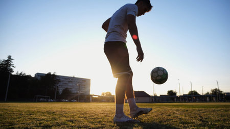 Professional footballer juggling soccer ball on stadium at sunset. Young man kicking ball at green field. Sportsman practicing tricks at meadow with sunlight at background. Freestyle football conceptの写真素材