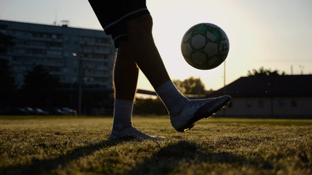 Legs of young man kicking ball at green field. Male feet of professional footballer juggling soccer ball on stadium at sunset. Sportsman practicing tricks outdoor. Concept of a freestyle footballの写真素材