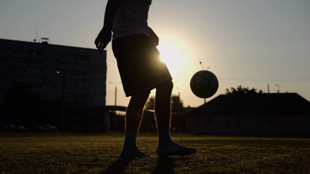 Legs of young man kicking ball at green field. Male feet of professional footballer juggling soccer ball on stadium at sunset. Sportsman practicing tricks outdoor. Concept of a freestyle footballの写真素材