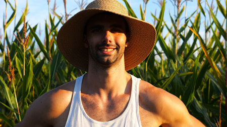 Happy smiling male farmer looks into camera standing near corn meadow. Portrait of young handsome agronomist in straw hat with maize field at background. Agricultural business conceptの写真素材