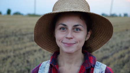 Happy smiling female farmer looks into camera standing at wheat field. Portrait of young beautiful agronomist in straw hat with barley meadow at background. Agricultural business conceptの写真素材