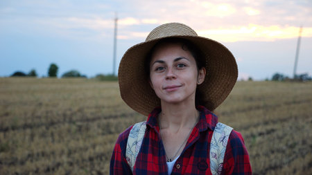 Happy smiling female farmer looks into camera standing at wheat field. Portrait of young beautiful agronomist in straw hat with barley meadow at background. Agricultural business conceptの写真素材