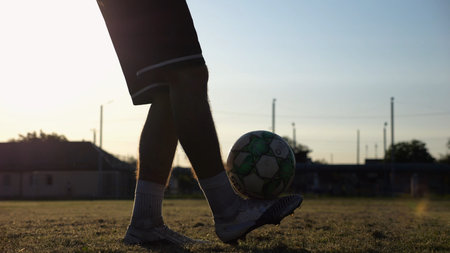 Legs of young man kicking ball at green field. Male feet of professional footballer juggling soccer ball on stadium at sunset. Sportsman practicing tricks outdoor. Concept of a freestyle footballの写真素材