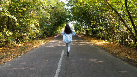 Young brunette woman running along road at forest on early autumn. Happy girl jogging among asphalt trail at nature. Unrecognizable female spending time at beautiful scenic. Close up Slow motionの写真素材