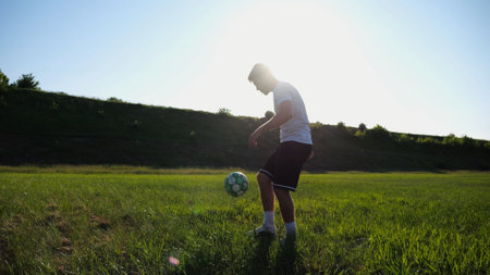 Sportsman juggling soccer ball on stadium at sunny day. Young man kicking ball at green field. Professional footballer practicing tricks at meadow with sunlight at background. Freestyle football.の写真素材