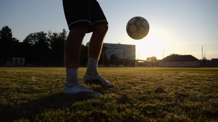 Legs of young man kicking ball at green field. Male feet of professional footballer juggling soccer ball on stadium at sunset. Sportsman practicing tricks outdoor. Concept of a freestyle footballの写真素材