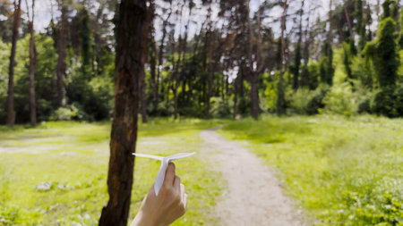 Female hand playing with paper airplane at summer forest. Girl walking with toy plane at woodland on sunny day. Young woman having fun at nature. Concept of leisure outdoor. Close upの写真素材