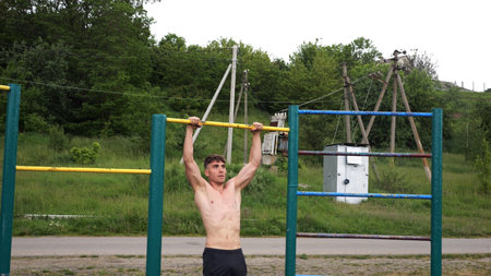 Muscular athlete doing pull ups on horizontal bar at sports ground. Young shirtless sportsman...