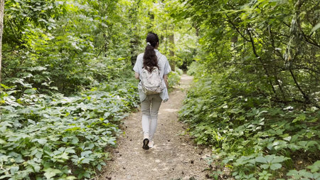 Young woman going along path at summer forest. Female with backpack walking among trail at wild...
