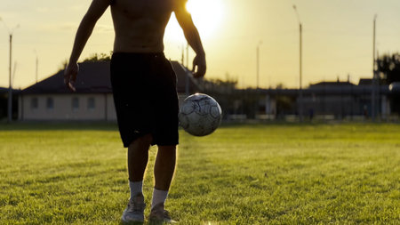 Male feet of professional footballer kicking ball at green field. Legs of young man juggling soccer...