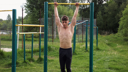 Muscular athlete doing pull ups on horizontal bar at sports ground. Young shirtless sportsman...