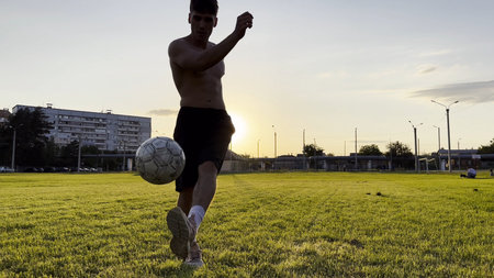 Young man juggling soccer ball on stadium at sunset. Professional footballer kicking ball at green field. Sportsman practicing tricks at meadow with sunlight at background. Freestyle footballの写真素材
