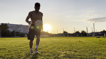 Young man juggling soccer ball on stadium at sunset. Professional footballer kicking ball at green field. Sportsman practicing tricks at meadow with sunlight at background. Freestyle footballの写真素材
