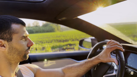 Profile of young handsome man driving a modern electric car with sunlight at background. Young male driver riding on electrical vehicle at countryside. Concept of journey and tripの写真素材