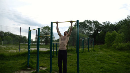 Muscular athlete doing pull ups on horizontal bar at sports ground. Young shirtless sportsman...