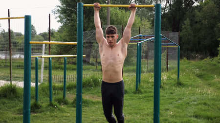 Muscular athlete doing pull ups on horizontal bar at sports ground. Young shirtless sportsman...