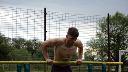 Male shirtless athlete doing push ups on parallel bars at nature. Strong sporty man exercising at...