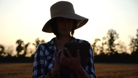Female agronomist in straw hat monitoring harvest on barley field at sunset. Adult farmer using...