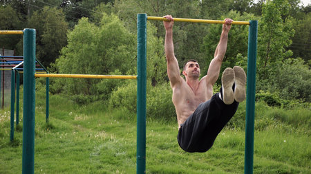 Athletic man trains abdominal muscles raising legs on horizontal bar at sports ground. Young...