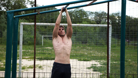 Young muscular sportsman doing pull ups at sports ground. Athletic guy exercising outdoor. Strong...