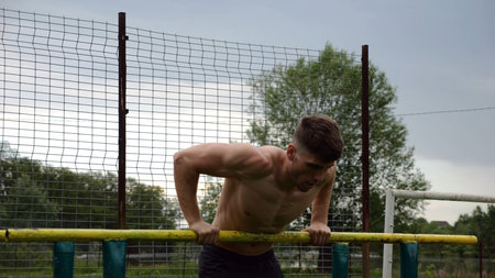 Male shirtless athlete doing push ups on parallel bars at nature. Strong sporty man exercising at...