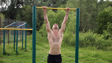 Muscular athlete doing pull ups on horizontal bar at sports ground. Young shirtless sportsman...